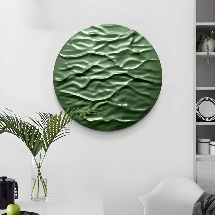 A modern interior features the Orenda Ripple Wave Green Wood Relief 3D Wall Art by Giant Sculptures, with its wavy pattern. Below, a table with a glass vase of palm leaves rests against minimalist white walls and shelves, completing the decor.