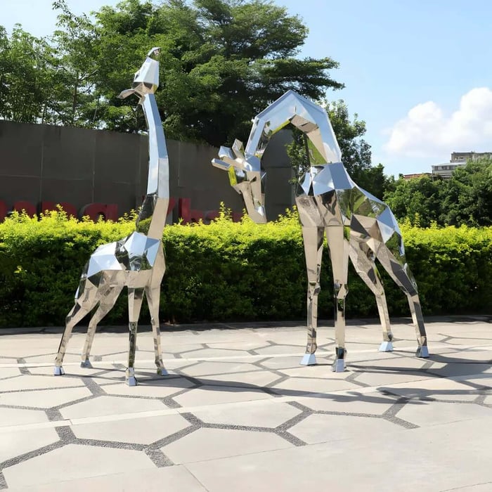 Pair of tall geometric stainless steel giraffe sculptures standing on a patterned stone plaza with greenery behind.