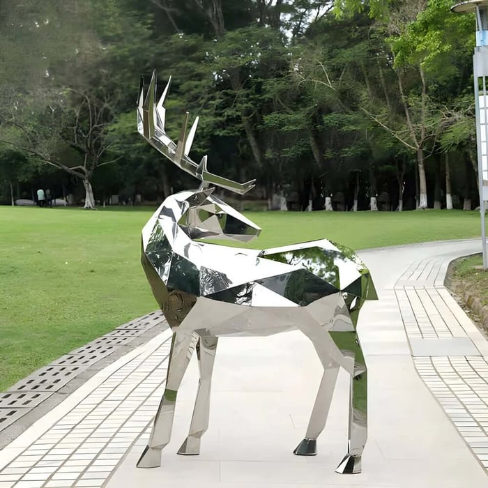 Mirror-finish geometric stag sculpture with antlers on a park walkway beside lawn and trees.