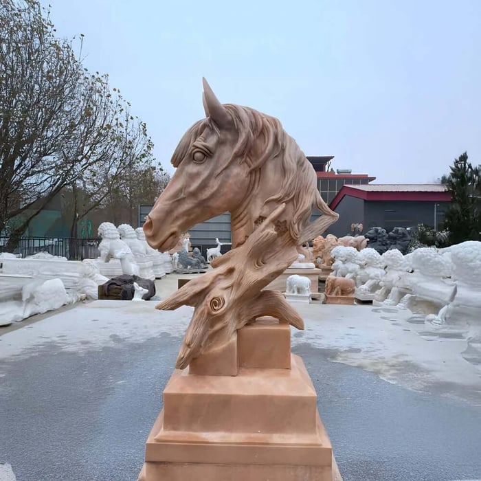 Sandstone horse head bust with flowing mane on a stepped pedestal in an outdoor sculpture yard surrounded by other stone statues.