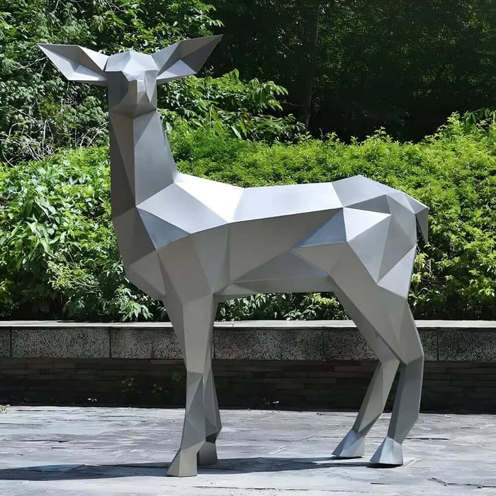 Matte grey, polygonal metal deer sculpture standing on a stone terrace, framed by dense green shrubs in an outdoor garden.