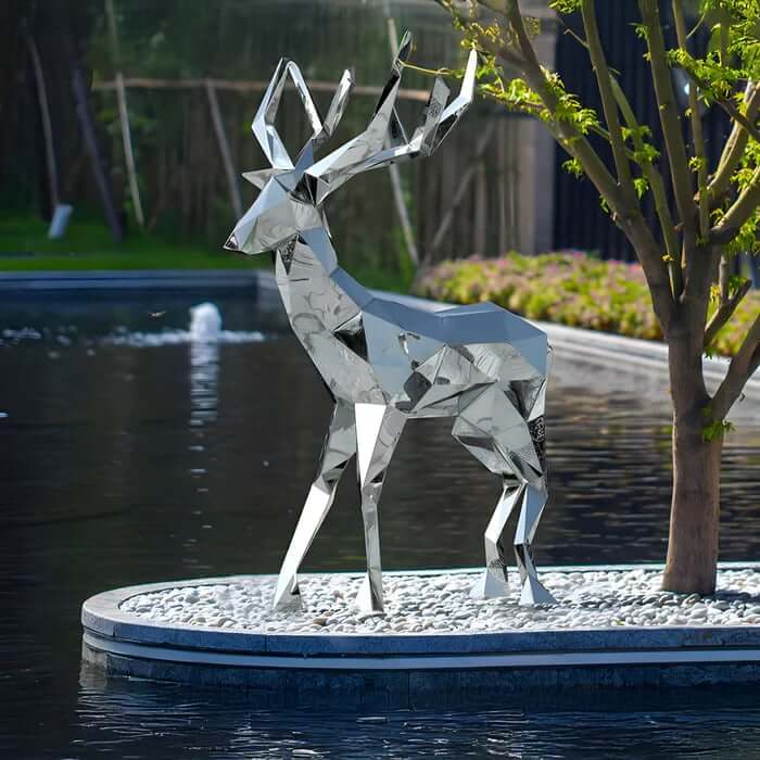 Faceted stainless-steel deer sculpture on a pebble island beside a reflective pond in a landscaped garden.
