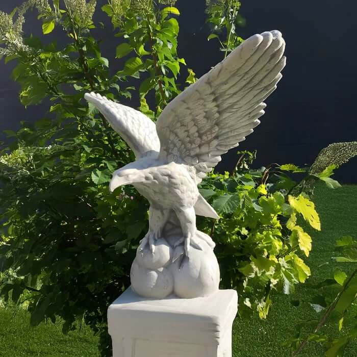 White stone eagle sculpture with wings spread, perched on a spherical base atop a pedestal in a leafy garden.