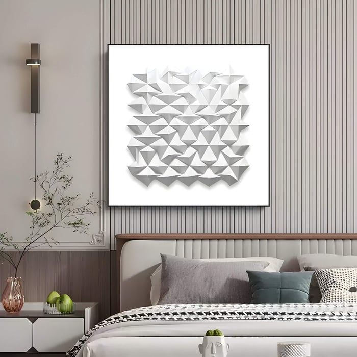 A modern bedroom in neutral tones showcases Giant Sculptures Geometrica Starburst White 3D Wood Relief Wall Art featuring folded shapes. The room includes a bed with a gray headboard, assorted pillows, a side table with flowers, and a decorative white light fixture on the wall.