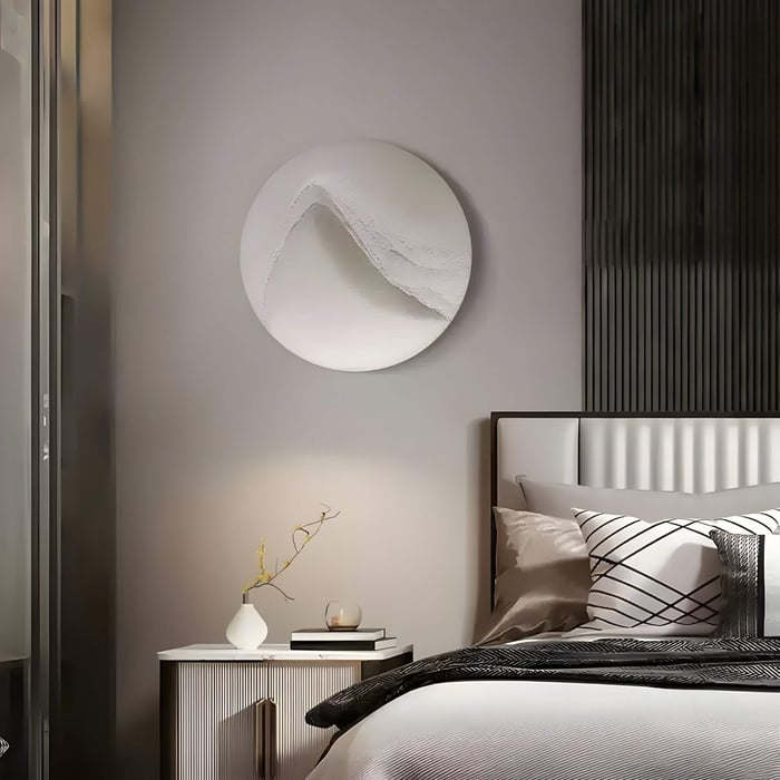 A round white wall sculpture with a textured ridge hangs above a modern nightstand in a serene, minimalist bedroom.