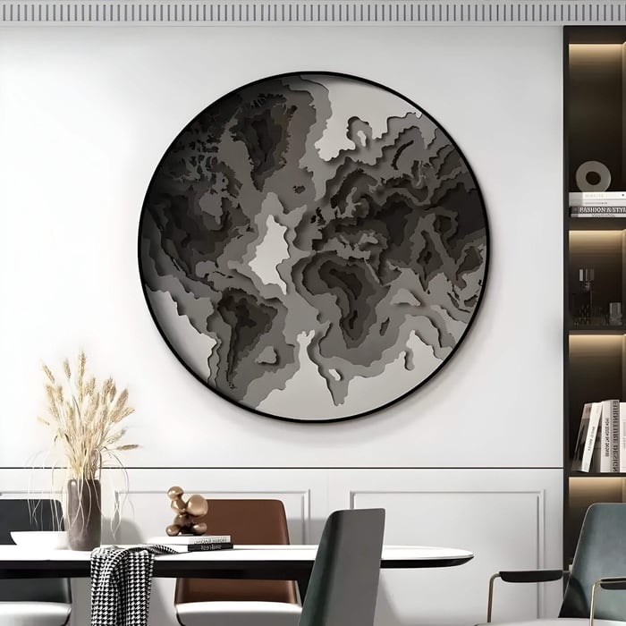 The Terranova Ash Grey Round Wood Carving 3D Wall Art by Giant Sculptures, with its textured sophistication, features an abstract world map. It complements a modern dining table adorned with wheat and nearby bookshelves in black, gray, and white tones.
