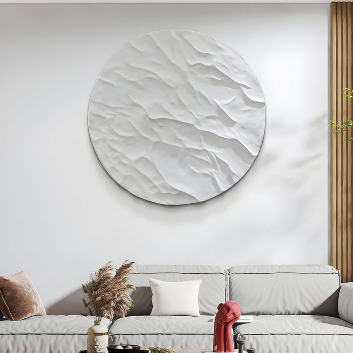 Large round white wall sculpture mimicking moon craters above a beige sofa with pampas grass in a calming, neutral setting.