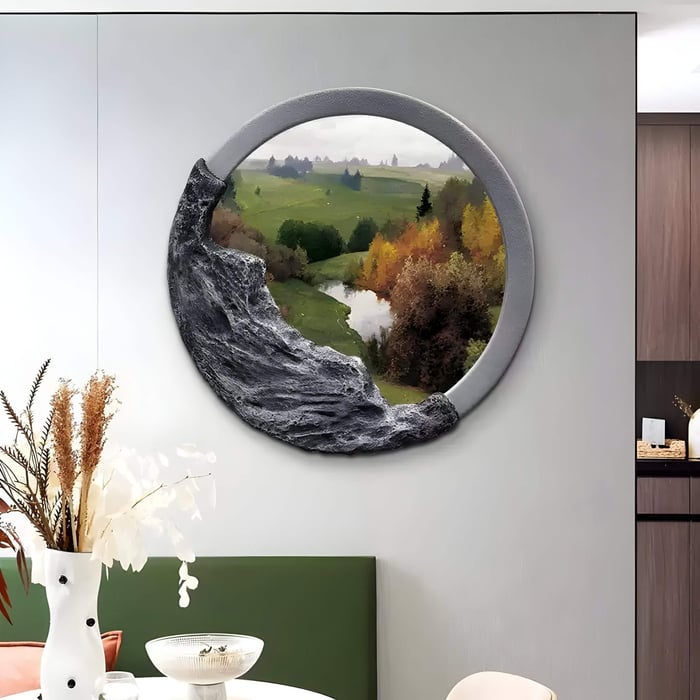 A round mirror with a sculpted gray stone frame reflects a scenic view similar to the Sylvara Meadow Vista Hand-Painted Wood Relief 3D Wall Art by Giant Sculptures, featuring lush greenery, a winding river, and autumn trees. Its mounted on a light gray wall above a green bench and table with a white vase and dried flowers.
