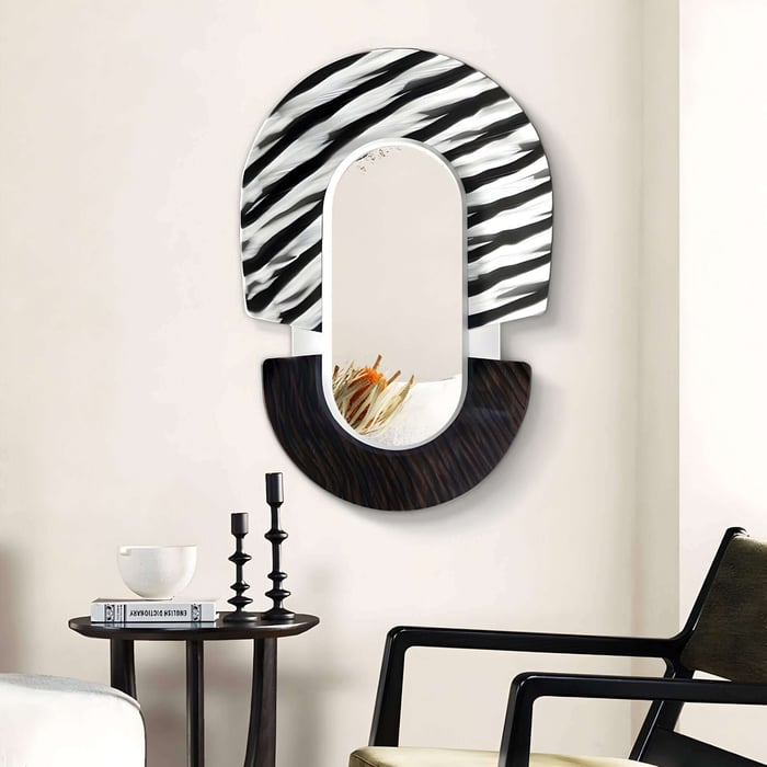 The Arcus Black & White Zebra Stripes Mirror 3D Wooden Wall Art by Giant Sculptures features a semicircular design with zebra stripes on top and a textured bottom. Below, a small shelf holds decorative dried grasses. Nearby, theres a table with candles and a bowl, plus a black chair in view.