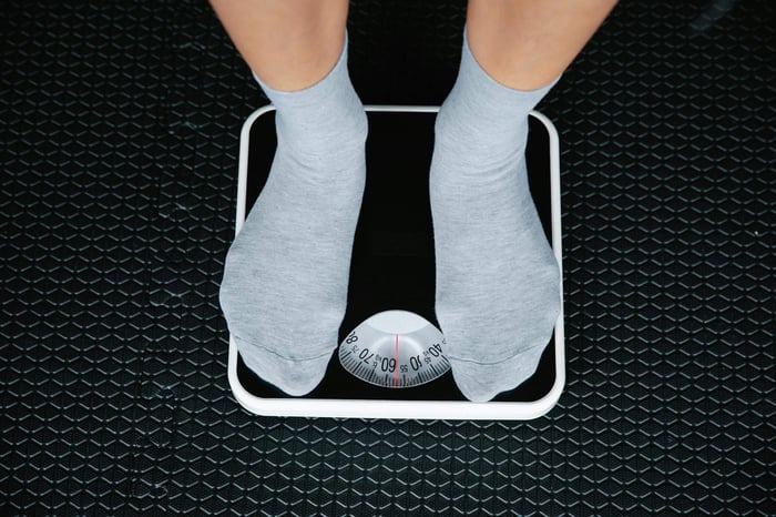 a person stepping on a scale probably to know if they have Extra Body Weight by knowing the number on the scale.  this person is wearing grey socks and is on a black scale on a black floor.