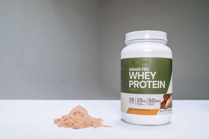MyFitFoods new Grass-Fed Whey Protein sits on a table in an empty environment, both the container and the power itself are shown.