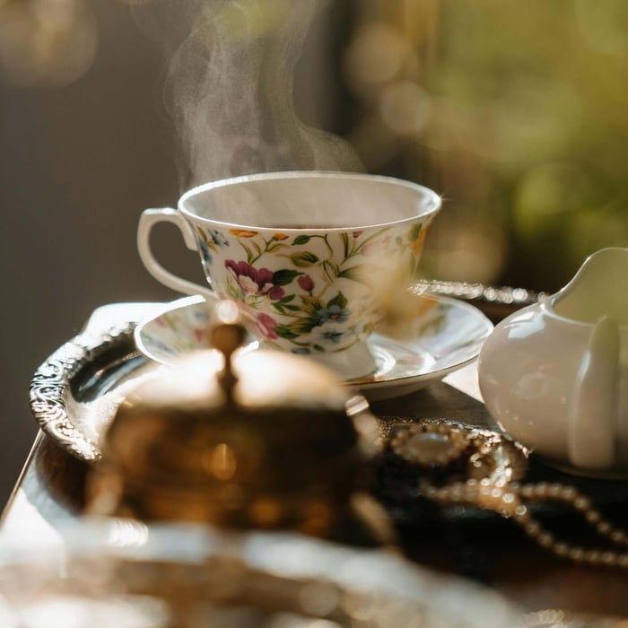 A very ornate cup and saucer presumably filled with tea sits in a blurry space signaling some elegant interior with natural light filling the space.