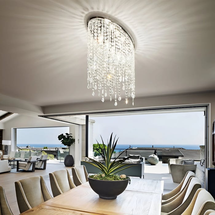 Bright, open-plan dining room with a ceiling-mounted chandelier adorned with cascading crystals, creating a luxurious effect. The room has large windows that open to an outdoor view with a garden and ocean.