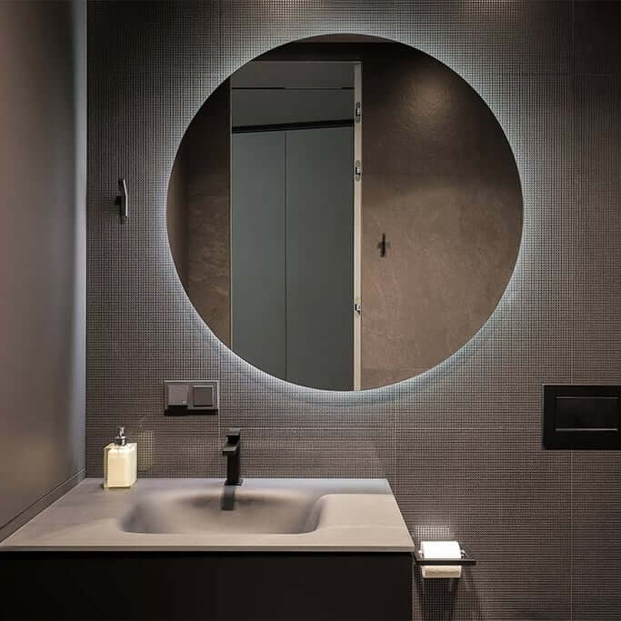 Round backlit bathroom mirror above a dark vanity with minimalist tap and textured wall tiles.