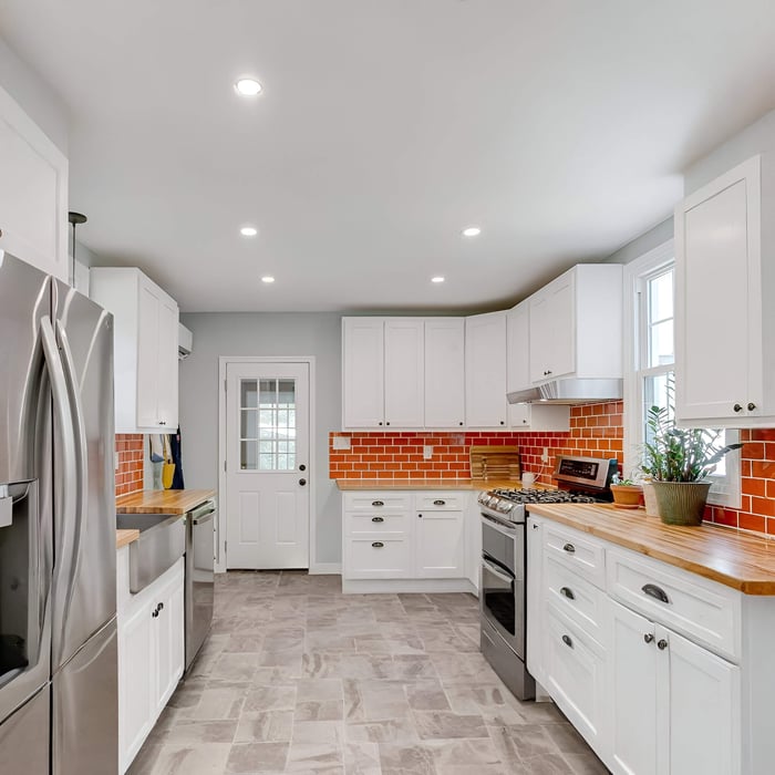 Narrow kitchen with white cabinets, orange subway-tile splashback, timber worktops, and recessed ceiling downlights.