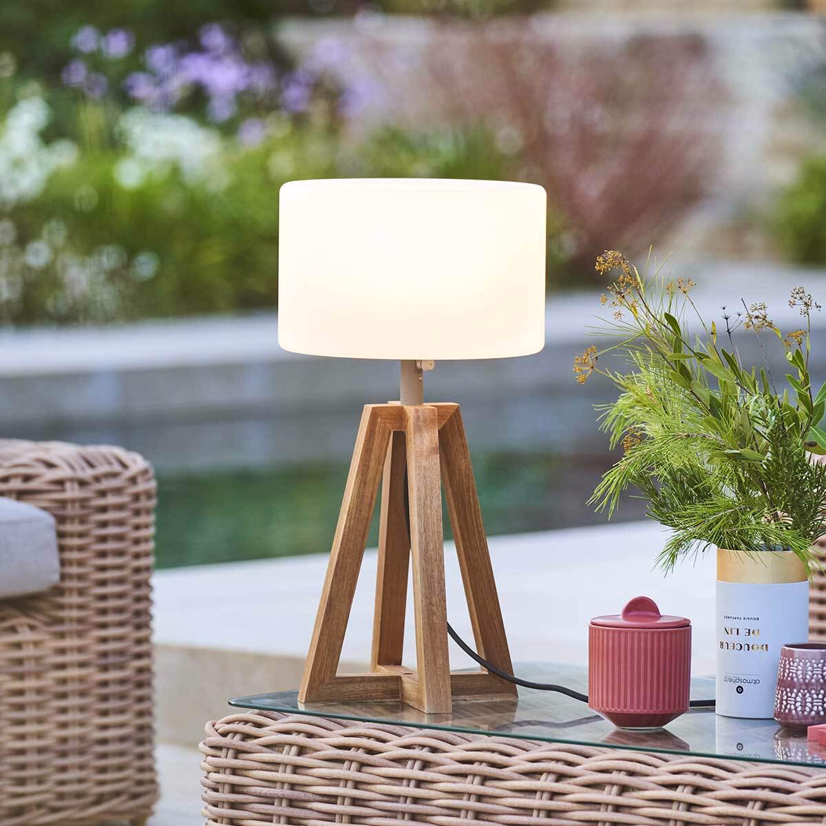 Outdoor table lamp with a warm white shade on a balcony dining table beside wine glasses, overlooking hills and city lights at dusk.