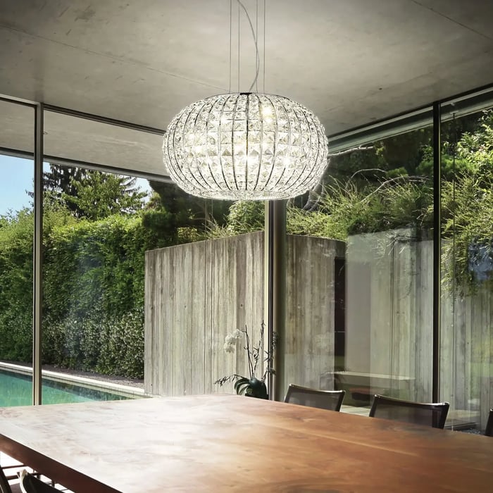 Modern dining space with a round glass chandelier above a wooden table, surrounded by floor-to-ceiling windows and greenery.