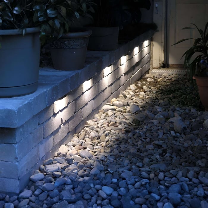 Low brick retaining wall with small recessed brick lights washing light onto a gravel border beside potted plants and an exterior door.