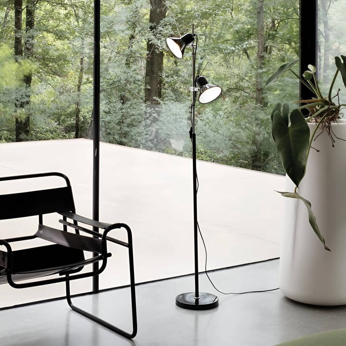 Double-head black industrial floor lamp by a glass wall and modern chair, overlooking a forest view.
