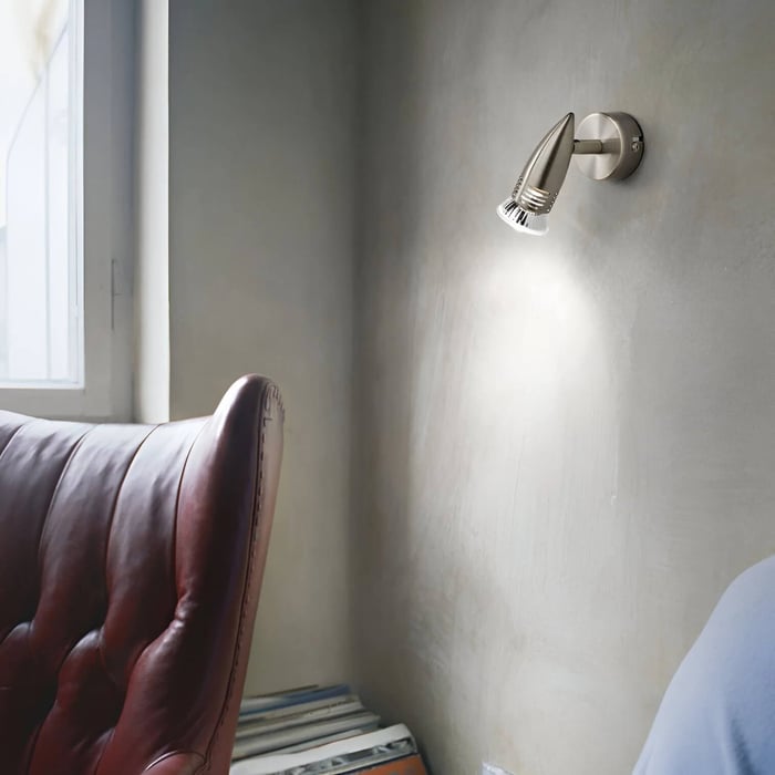 Close-up of a brushed-metal wall spotlight casting light beside a leather chair and window, ideal for task lighting.