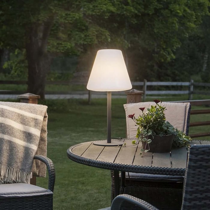 Outdoor table lamps with a white shade on a round garden table, surrounded by outdoor chairs and greenery at dusk.