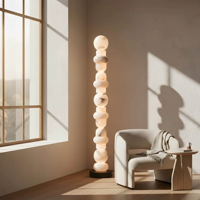 Sunlit corner with sculptural stacked alabaster floor lamp next to curved armchair and large window.