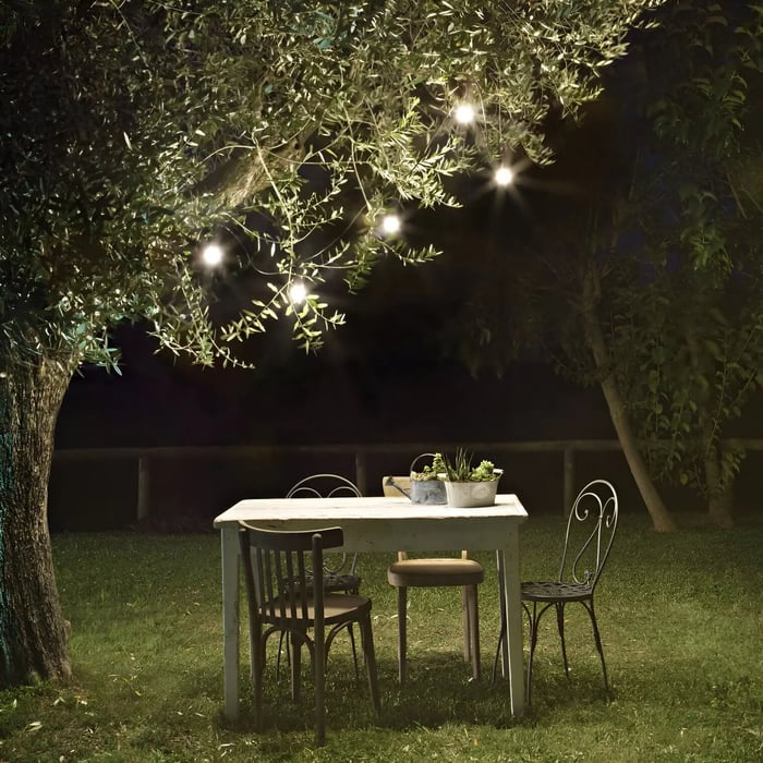 Soft outdoor string lights hanging through tree branches above a small bistro table and chairs on a lawn at night.
