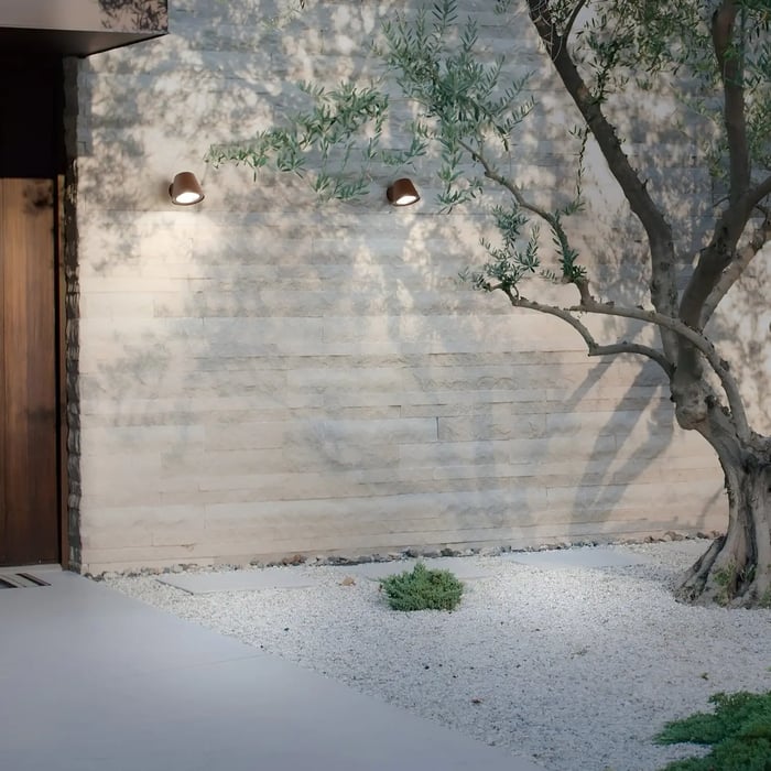 Minimalist home entrance with two small brown wall-mounted lights casting soft glow on textured stone wall beside an olive tree.