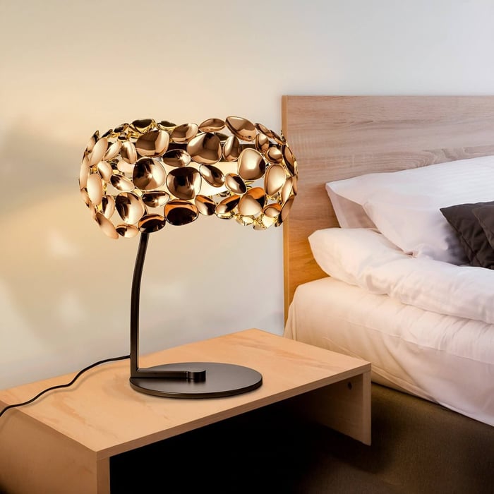 Close-up of a bedside table lamp with a sculptural gold metallic shade, placed next to a bed with white bedding and a wooden headboard.