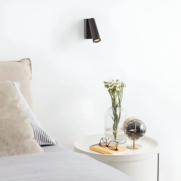 A minimal bedside setup with a sleek black wall-mounted sconce providing focused lighting. The side table holds a modern clock, glasses, and a vase with fresh white flowers, complementing the neutral tones of the bed.
