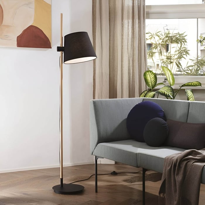 Modern floor lamp with black drum shade and wooden stand casting warm light beside a gray sofa.