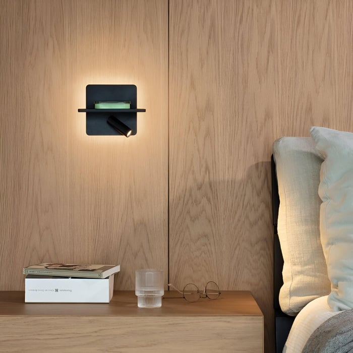 Contemporary bedroom with wooden wall panels, black wall lamp with integrated shelf, and bedside table with books, glass, and eyeglasses.