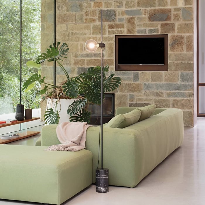 Slim floor lamp with exposed globe bulb and marble base in a stone-walled modern living room.