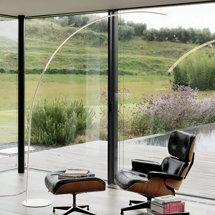  A sleek curved floor lamp next to a black lounge chair and ottoman, set in front of floor-to-ceiling glass windows overlooking a green outdoor space.