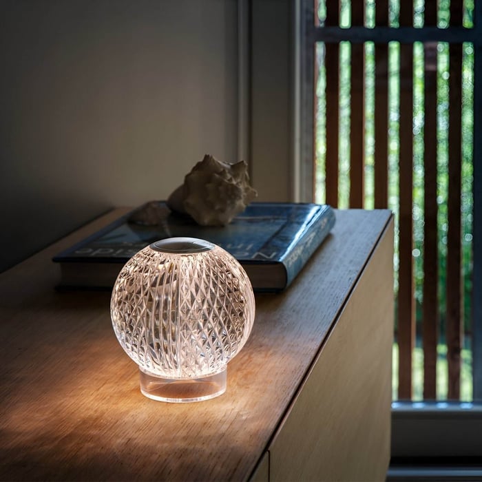 Small crystal orb table lamp glowing softly on a wooden surface beside books and seashell decor near a window.