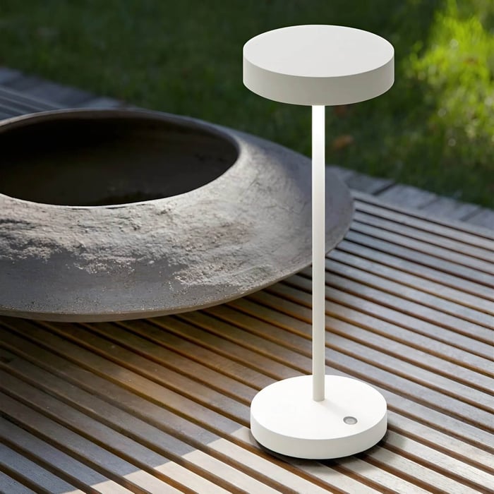 White modern outdoor table lamp on a wooden deck beside a large stone fire bowl, with a green lawn in the background.
