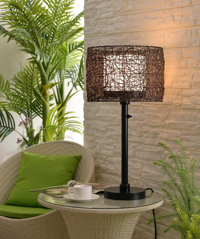 Woven rattan-style table lamp on a small round side table beside a wicker chair, set against a textured stone wall and plants.
