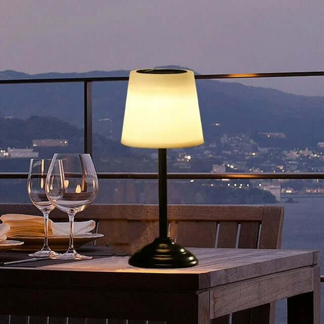 Outdoor table lamp with a warm white shade on a balcony dining table, beside wine glasses, overlooking a city and hills at dusk.