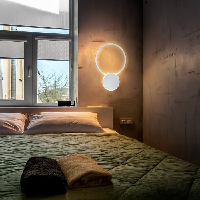 Industrial-style bedroom with a concrete wall, green bedding, and a glowing circular modern wall sconce beside the window.