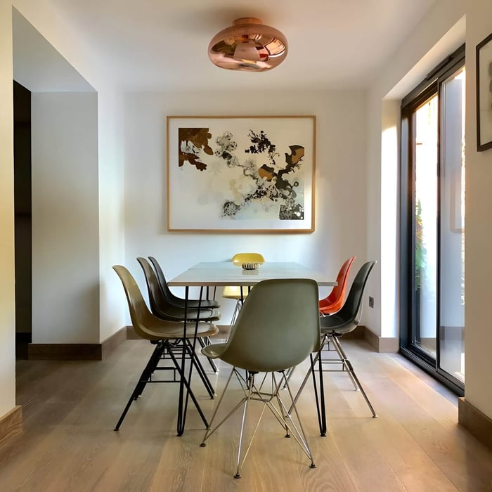 Contemporary dining nook with copper ceiling light, abstract wall art, and mismatched chairs in a sunlit corner near glass doors.