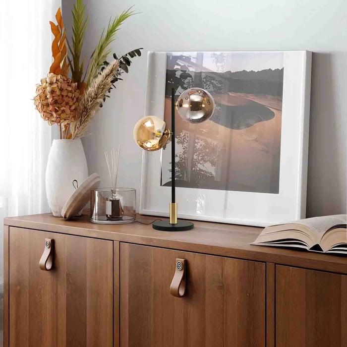 Slim black-and-brass modern table lamp with twin globe shades on a wooden sideboard beside framed art and dried florals.