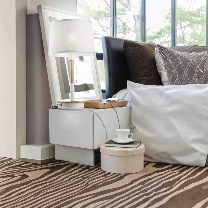White-shaded table lamp on a modern bedside cabinet next to a bed and large window.