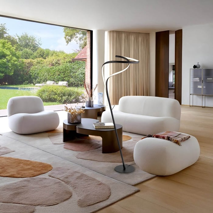 Bright minimal living room with floor-to-ceiling glass opening to a garden, white low-profile seating, round wooden tables, and a sculptural floor lamp.