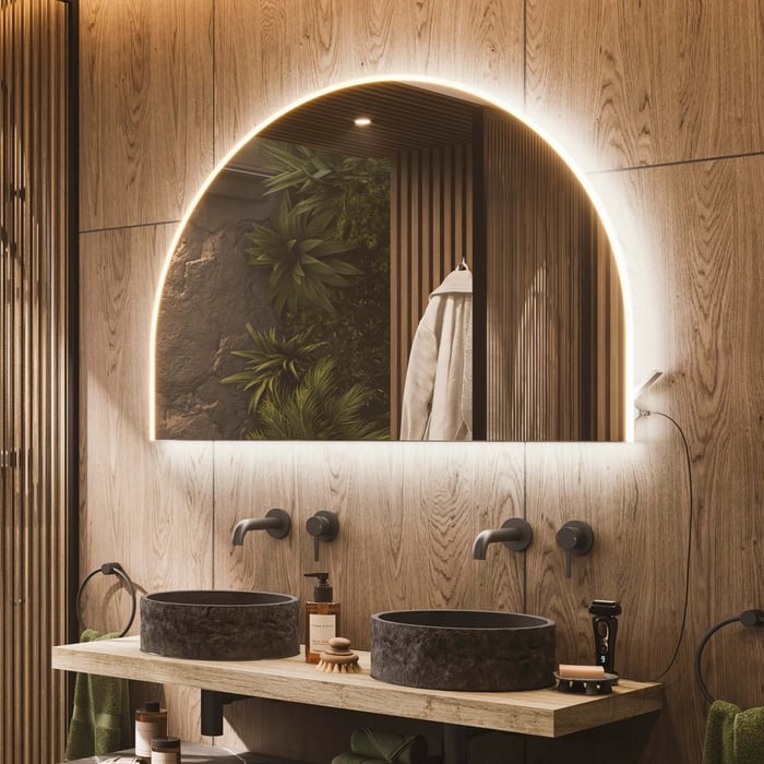 Warm wood bathroom with a large arch-shaped backlit mirror, dual stone sinks, and subtle recessed lighting.