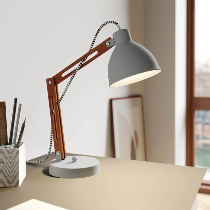 Grey and wood desk lamp with exposed cord on a tidy work surface in front of a sunlit window.