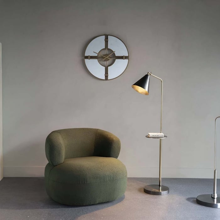 Slim brass-and-black industrial floor lamp with small tray next to a rounded green chair against a gray wall.