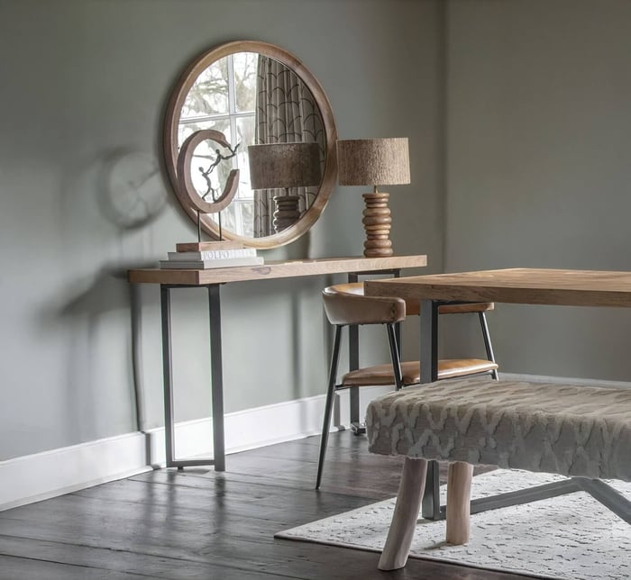Rustic wooden console table with matching lamp and round mirror, paired with a leather chair and bench in a neutral-toned room.