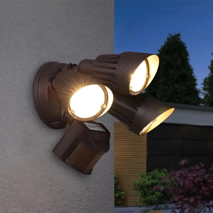 Three-head bronze security floodlight with a motion sensor lit up on an exterior wall at dusk, with a house and trees softly visible in the background.