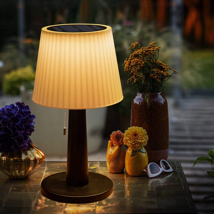 Solar-powered table lamp with a pleated shade emitting warm light, placed on an outdoor table surrounded by flowers and decor.