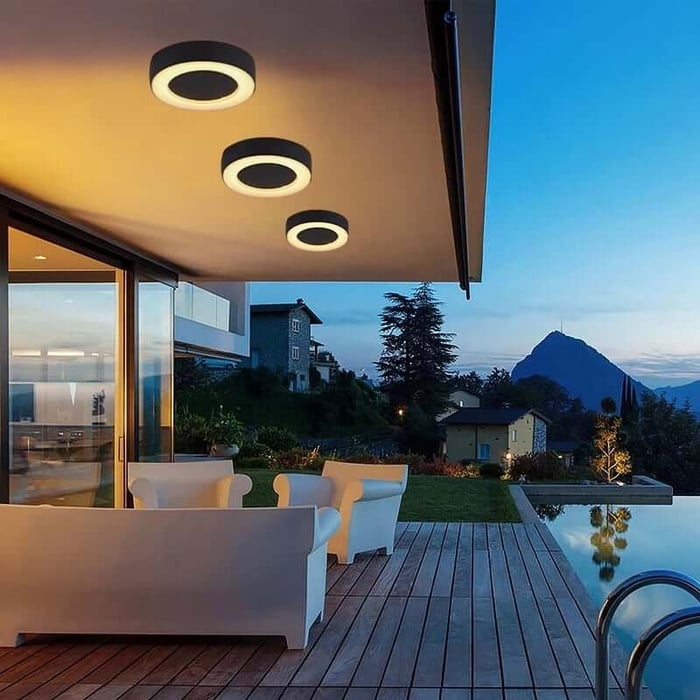Modern terrace beside a pool with circular outdoor ceiling lights mounted under the roof, overlooking a landscaped garden at dusk.